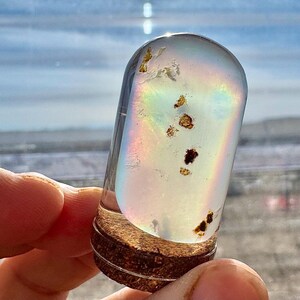 May include: A clear, cylindrical glass vial with a cork base, containing iridescent liquid and small, dark particles. The vial is held up against a bright, out-of-focus background, creating a rainbow effect within the glass.