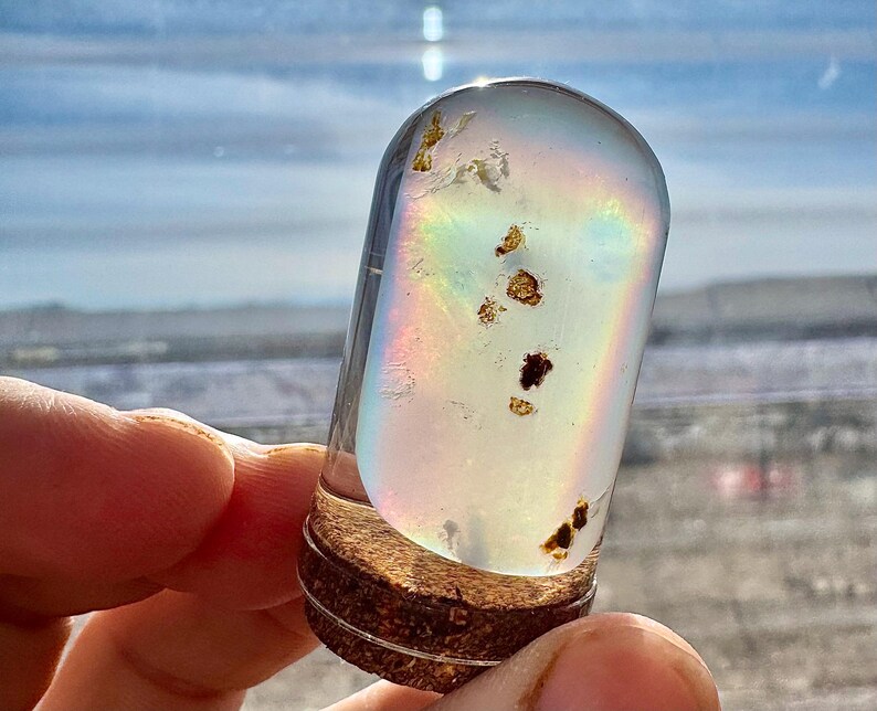 May include: A clear, cylindrical glass vial with a cork base, containing iridescent liquid and small, dark particles. The vial is held up against a bright, out-of-focus background, creating a rainbow effect within the glass.