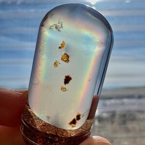 May include: A clear, cylindrical glass vial with a wooden base, containing iridescent light and small, dark inclusions. The vial is held in a hand, with a rainbow effect visible inside. The background is a bright, sunny sky.