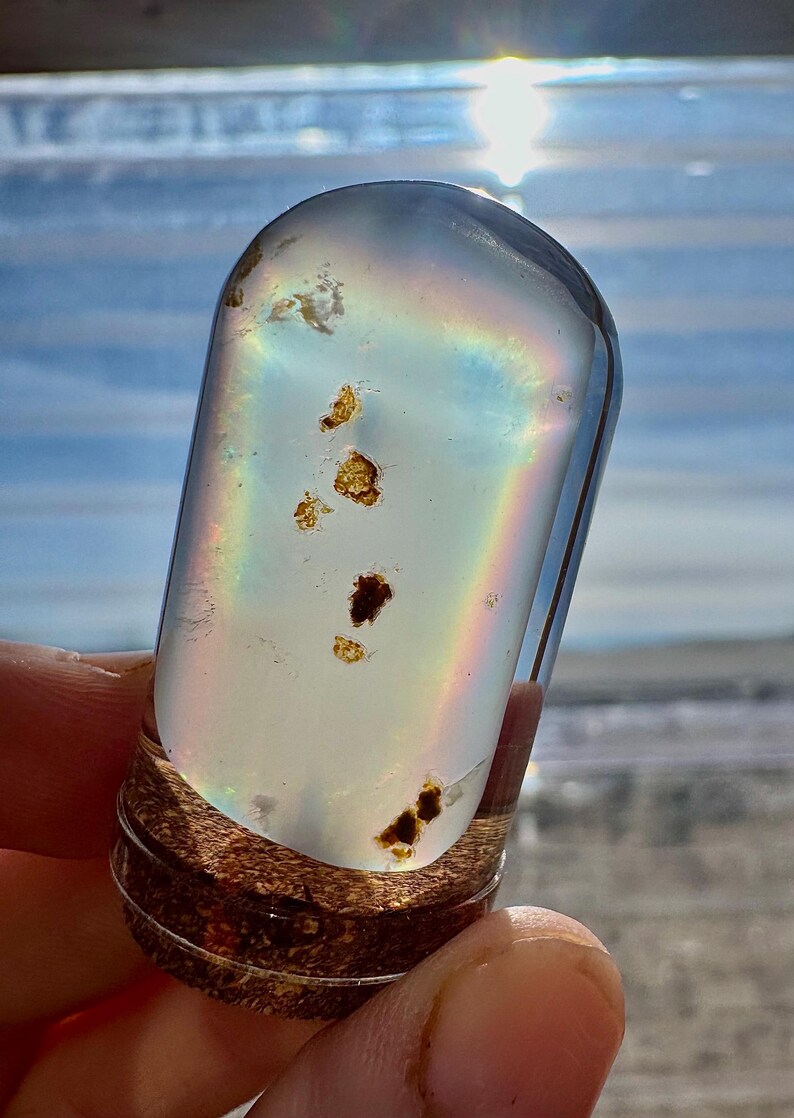 May include: A clear, cylindrical glass vial with a wooden base, containing iridescent light and small, dark inclusions. The vial is held in a hand, with a rainbow effect visible inside. The background is a bright, sunny sky.