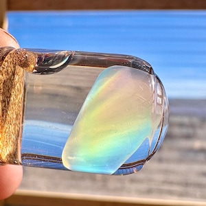 May include: A clear, rectangular pendant with a rainbow-colored, iridescent stone. The pendant is held up against a backdrop of a blue sky and water, creating a beautiful display of light and color.