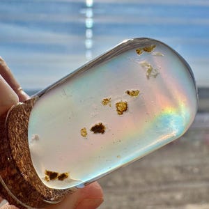 May include: A clear, teardrop-shaped glass bottle stopper with a wooden base. The glass contains small, brown inclusions and reflects a rainbow of colors. The stopper is held against a blurred background of a beach and sky.