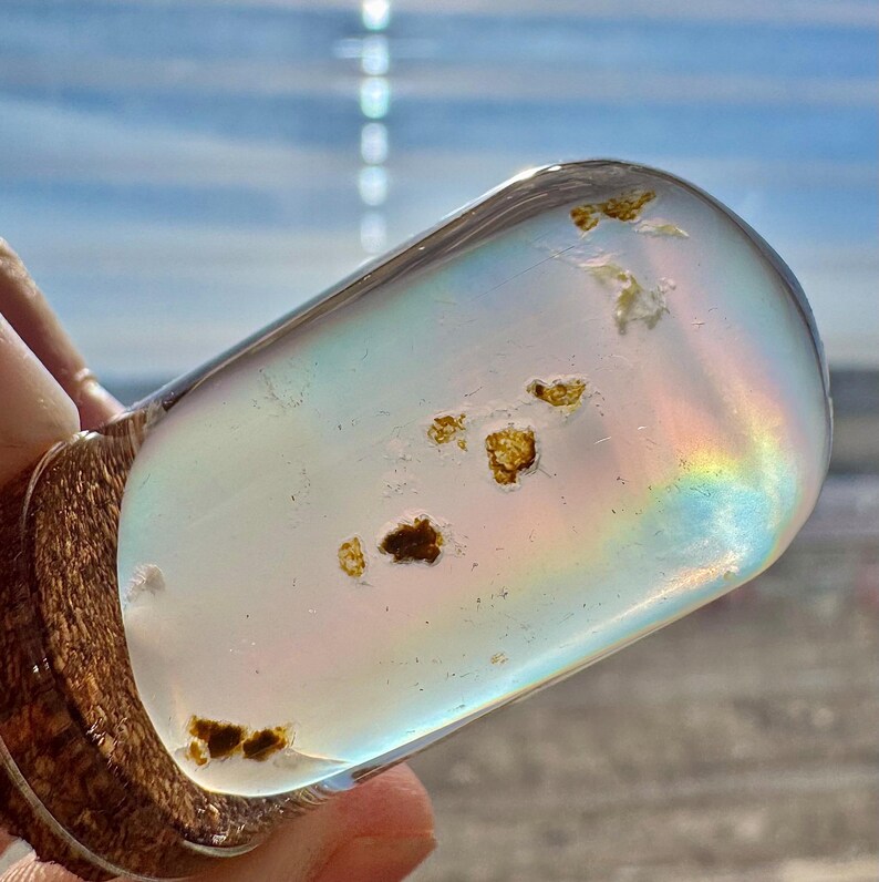 May include: A clear, teardrop-shaped glass bottle stopper with a wooden base. The glass contains small, brown inclusions and reflects a rainbow of colors. The stopper is held against a blurred background of a beach and sky.