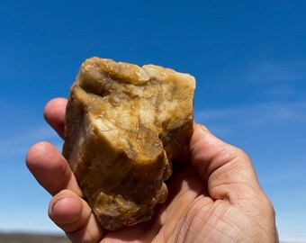 May include: A rough, natural stone with a mix of tan and cream colors. The stone has a layered texture and is held against a clear blue sky. The stone appears to be a mineral specimen.