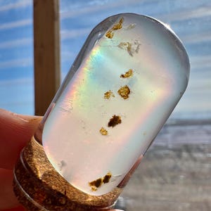May include: A clear, oval-shaped glass object with embedded golden-brown inclusions. The glass refracts light, creating a rainbow effect. The base is a textured, brown material. The object is held in a hand.