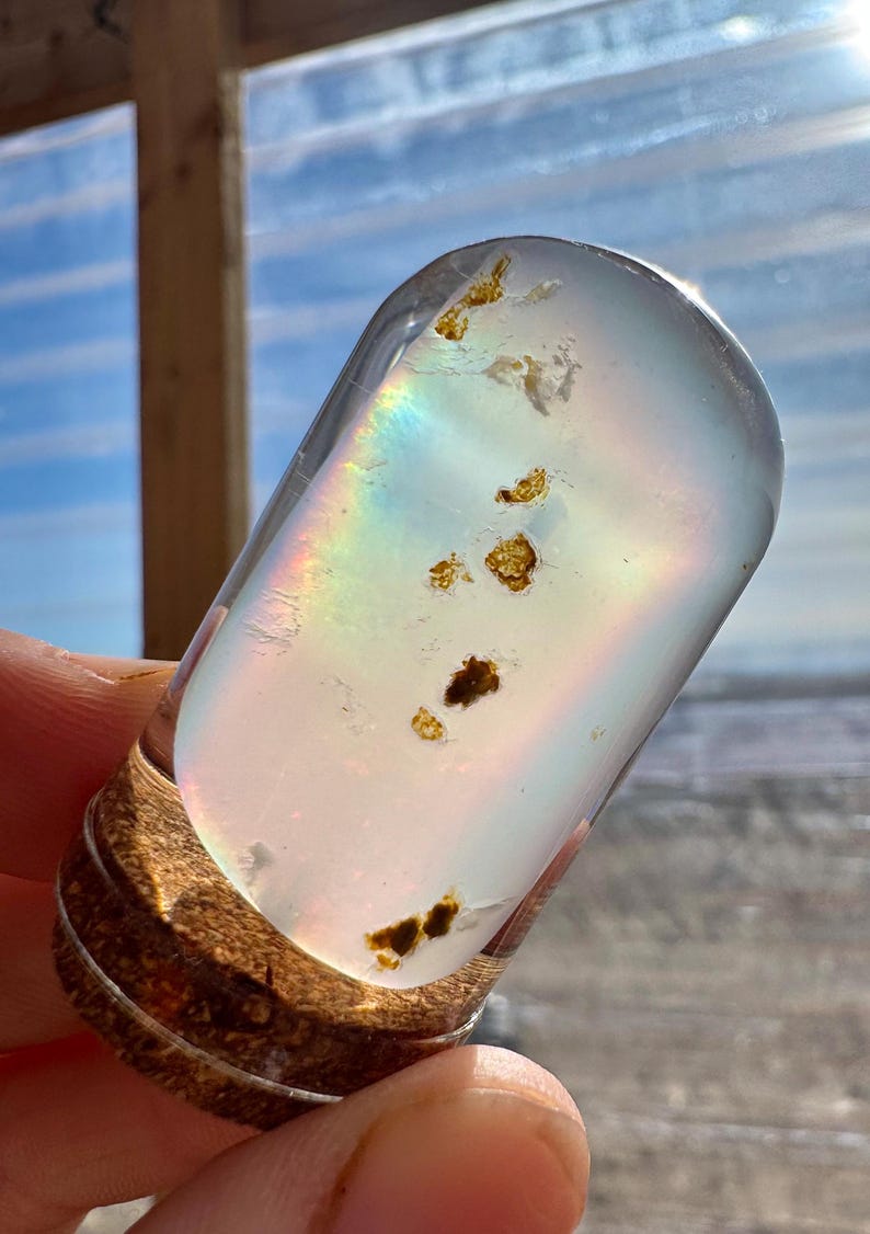 May include: A clear, oval-shaped glass object with embedded golden-brown inclusions. The glass refracts light, creating a rainbow effect. The base is a textured, brown material. The object is held in a hand.