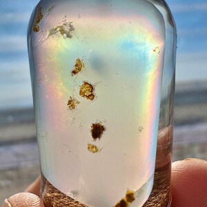 May include: A clear, cylindrical glass bottle with a cork base, filled with a pearlescent liquid and small, golden-brown inclusions. A rainbow effect is visible within the glass. The bottle is held against a bright, sunny background.