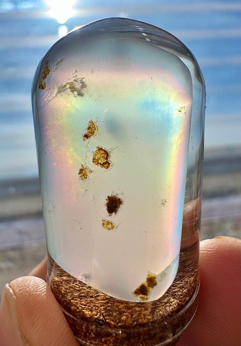 May include: A clear, cylindrical glass bottle with a cork base, filled with a pearlescent liquid and small, golden-brown inclusions. A rainbow effect is visible within the glass. The bottle is held against a bright, sunny background.