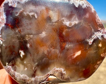 May include: A polished, oval-shaped stone with a mix of colors. The stone features a blend of brown, red, and white hues, with a translucent quality. The surface has a smooth, glossy finish, and the edges show a natural, rough texture.