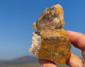 May include: A rough-hewn, natural stone with a mix of colors, including tan, brown, and hints of purple. The rock has a layered appearance and a slightly rough texture. The background features a clear blue sky and a distant, blurred landscape.