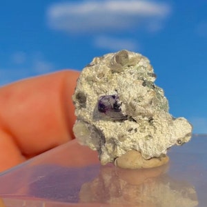 May include: A small, rough-textured rock formation with a prominent purple crystal. The rock has a light gray color with hints of silver and sits on a clear surface against a blue sky background.