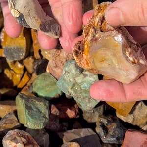 May include: A collection of colorful rocks and stones, including shades of green, brown, and tan. The image shows a variety of shapes and textures, with some stones held in a person's hand. The rocks appear to be natural and unpolished.