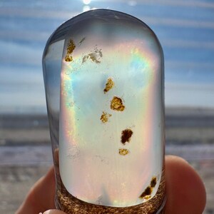 May include: A clear, rounded glass object with internal inclusions and a rainbow iridescence. The base has a textured, brown appearance. The object is held in a hand, with a bright sky in the background.