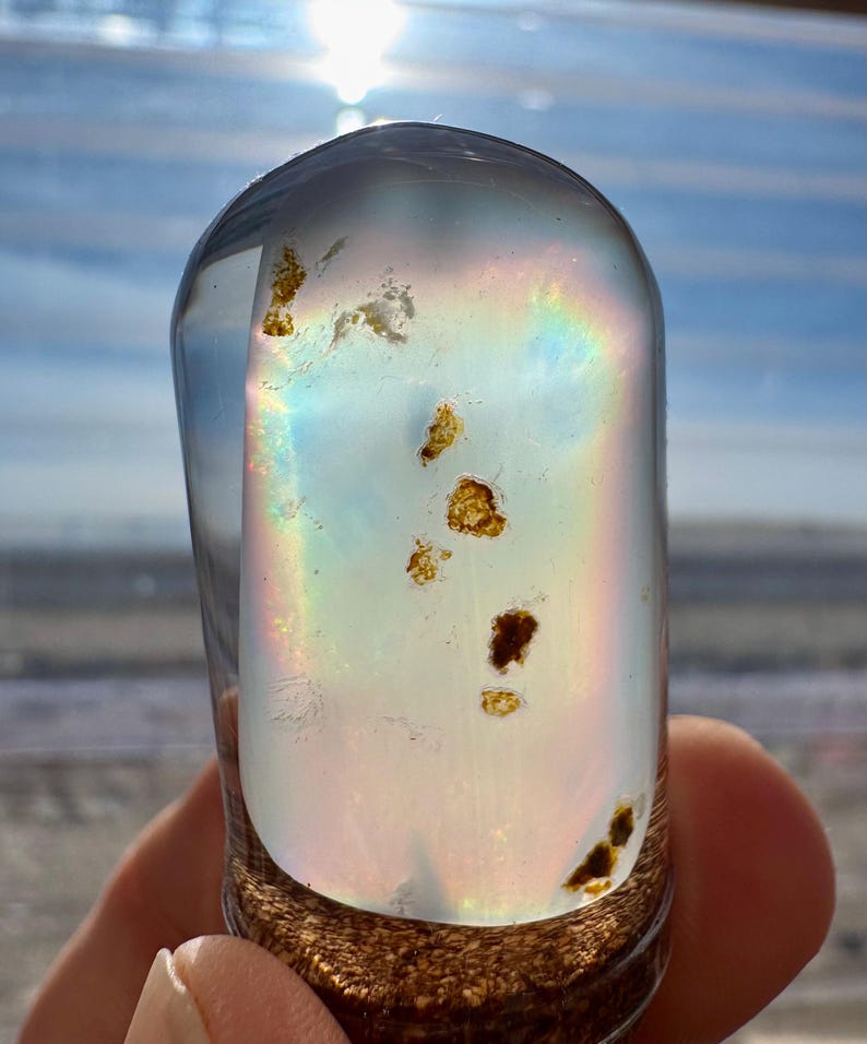 May include: A clear, rounded glass object with internal inclusions and a rainbow iridescence. The base has a textured, brown appearance. The object is held in a hand, with a bright sky in the background.