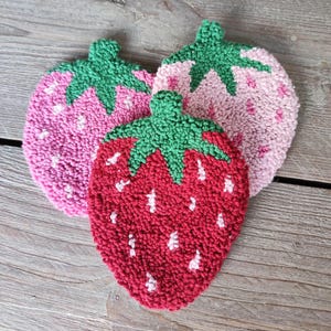 May include: Three handmade felt strawberry coasters. One is red, one is pink, and one is light pink. The strawberries have green leaves and white seeds.