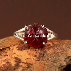 Op de afbeelding: Een zilveren ring met een gegraveerde rode edelsteen. De ring ligt op een bruine achtergrond. "Silver Artisan Jewels" staat op de ringband.