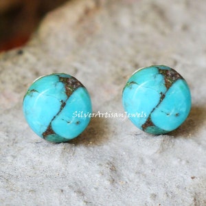 May include: A pair of round turquoise earrings. Each earring features a vibrant blue-green color with brown veining. The earrings are set against a neutral background. The text "Silver Artisan Jewels" is visible on the image.