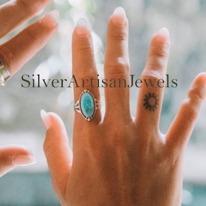 May include: A silver ring with a turquoise oval stone. The ring is on a person's finger, which also has a small sun tattoo.