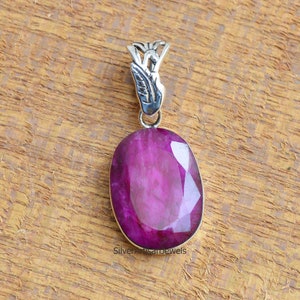 Puede incluir: Un colgante de plata con una piedra preciosa grande, ovalada, facetada y púrpura. El colgante tiene una baíl ornada y detallada.