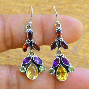 Può includere: Orecchini pendenti in argento sterling con una pietra preziosa gialla a forma di lacrima circondata da pietre preziose viola e verdi. La parte superiore degli orecchini presenta un gruppo di pietre preziose marroni.