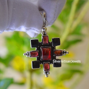 May include: A silver pendant with a geometric design featuring red and black gemstones. The pendant is in the shape of a cross with a silver bail.