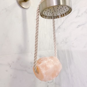May include: A pink Himalayan salt rock hanging from a shower head with a white rope. The shower head is silver and is spraying water.