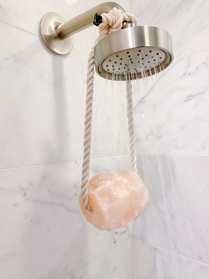 May include: A pink Himalayan salt rock hanging from a shower head with a white rope. The shower head is silver and is spraying water.