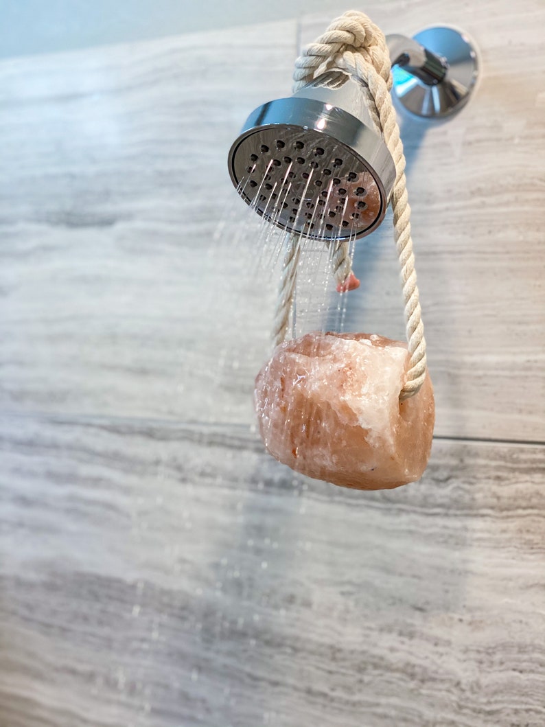 May include: A pink Himalayan salt rock hanging from a shower head with a rope. Water is flowing from the shower head.