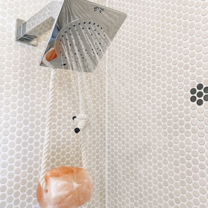 May include: A white rope hangs from a silver shower head with a pink Himalayan salt rock attached to the end. The shower head is mounted on a white tiled wall.