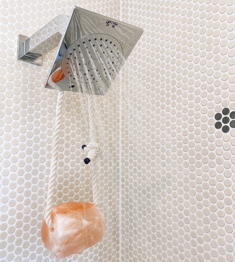 May include: A white rope hangs from a silver shower head with a pink Himalayan salt rock attached to the end. The shower head is mounted on a white tiled wall.