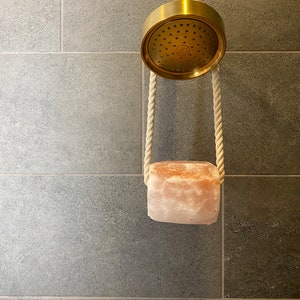 May include: A gold shower head with a pink Himalayan salt rock hanging from it with white rope.