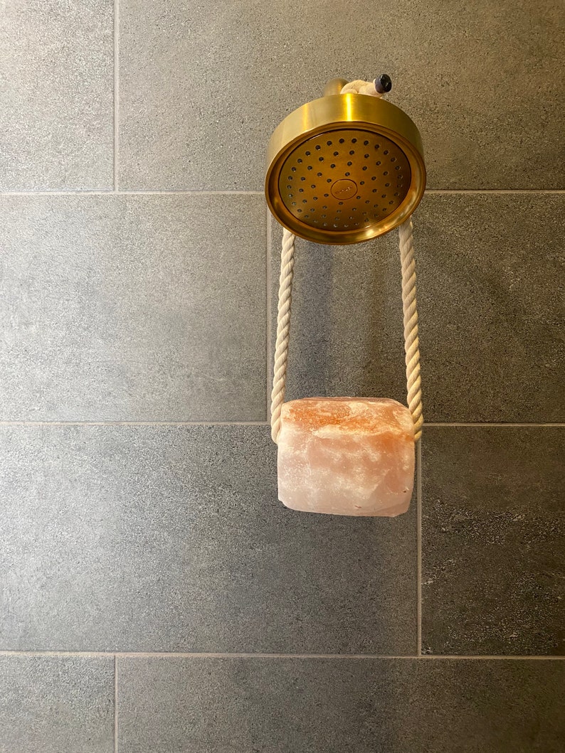 May include: A gold shower head with a pink Himalayan salt rock hanging from it with white rope.