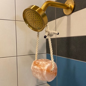 May include: A gold shower head with a white rope hanging from it. A large pink Himalayan salt rock is attached to the rope.