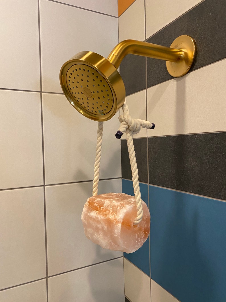 May include: A gold shower head with a white rope hanging from it. A large pink Himalayan salt rock is attached to the rope.