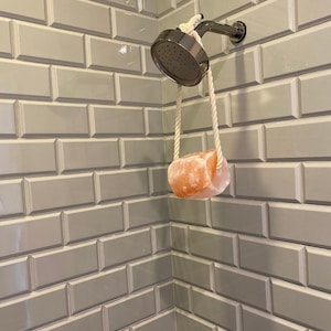 May include: A white rope shower head holder with a pink Himalayan salt soap bar hanging from it. The shower head is silver and the soap bar is hanging in a white tiled shower.