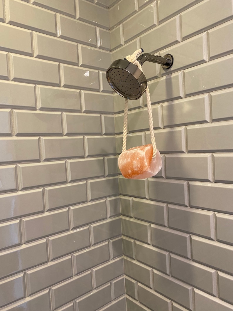 May include: A white rope shower head holder with a pink Himalayan salt soap bar hanging from it. The shower head is silver and the soap bar is hanging in a white tiled shower.