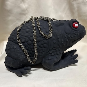 Könnte beinhalten: Eine mattschwarze, krötenförmige Tasche mit strukturierter, noppiger Oberfläche und leuchtend roten Augen mit weißen Pupillen. Die Tasche verfügt über einen langen, metallischen Kettenriemen aus ineinandergreifenden Ringen auf dem Rücken.