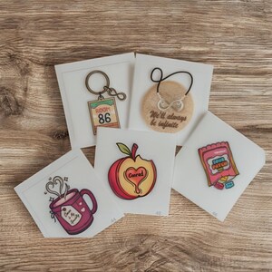 May include: A collection of stickers on a wooden surface. The stickers include a keychain with "Room 86", a cookie with "We'll always be infinite", a red apple with "Conrad", a mug with "Coffee is my Soulmate", and a Sour Patch Kids candy.