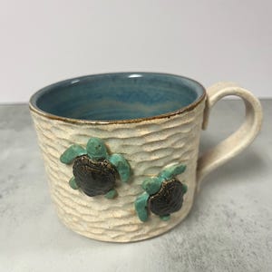Puede incluir: Taza de cerámica color crema con exterior texturizado e interior azul claro. Dos tortugas marinas en 3D están adheridas al exterior de la taza. El asa es curvada. Adecuada para bebidas frías o calientes.