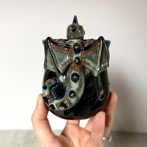 May include: A black ceramic mug with a dragon design. The dragon has outstretched wings, a spiked head, and a tail that wraps around the mug. The wings and dragon details have a blue and brown glaze.