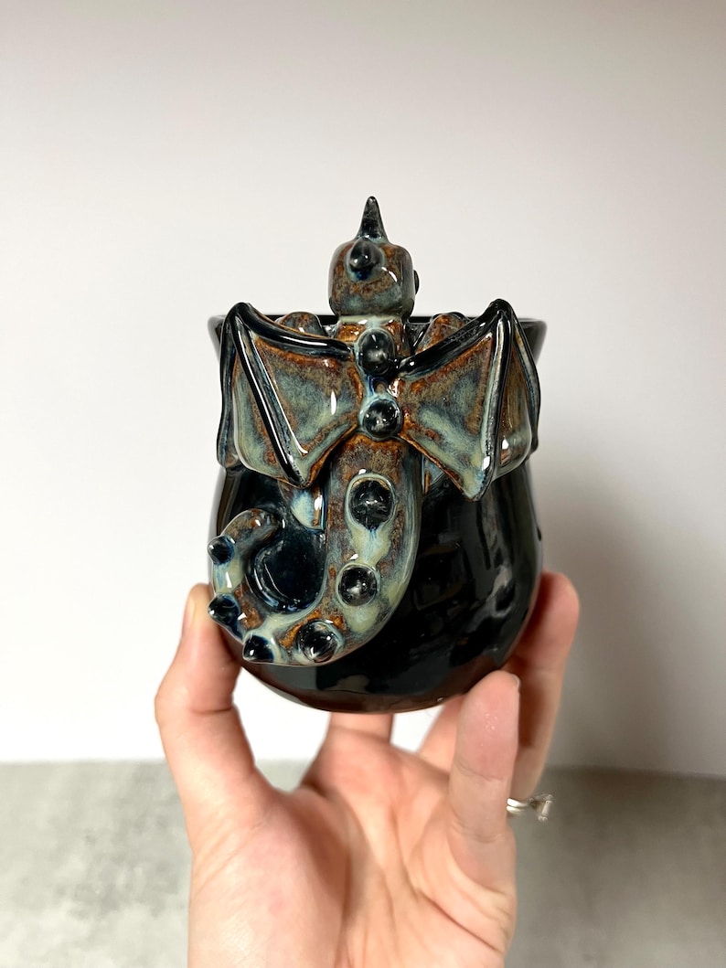 May include: A black ceramic mug with a dragon design. The dragon has outstretched wings, a spiked head, and a tail that wraps around the mug. The wings and dragon details have a blue and brown glaze.