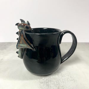 May include: Black ceramic mug with a dragon figurine attached to the side. The dragon has brown, blue, and green accents. The mug has a glossy finish and a curved handle. The mug is designed for beverages.