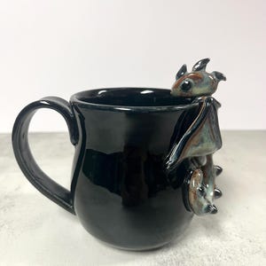 May include: Black ceramic mug with a glossy finish. A detailed, three-dimensional bat figure is attached to the side of the mug. The bat has brown, blue, and black coloring. The mug has a curved handle.