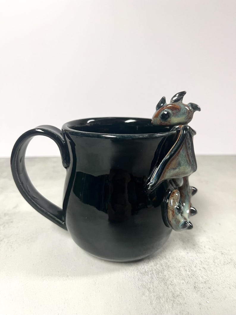 May include: Black ceramic mug with a glossy finish. A detailed, three-dimensional bat figure is attached to the side of the mug. The bat has brown, blue, and black coloring. The mug has a curved handle.