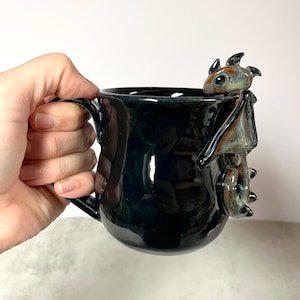 May include: Black ceramic mug with a glossy finish, featuring a detailed dragon sculpture on the side. The dragon has brown, blue, and green accents. The mug has a handle for easy gripping and is suitable for hot or cold beverages.