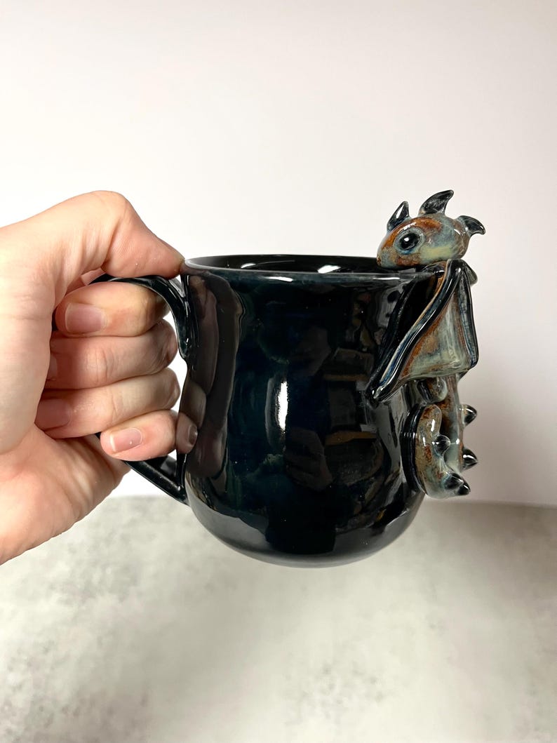 May include: Black ceramic mug with a glossy finish, featuring a detailed dragon sculpture on the side. The dragon has brown, blue, and green accents. The mug has a handle for easy gripping and is suitable for hot or cold beverages.