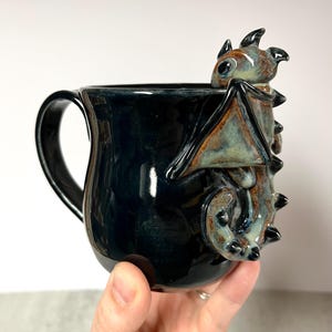 May include: A black ceramic mug with a dragon sculpture on the side. The dragon is brown, blue, and tan. The mug has a handle and is held by a hand.