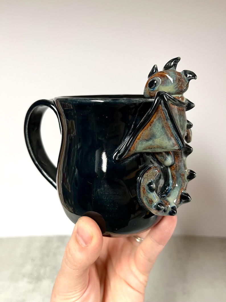 May include: A black ceramic mug with a dragon sculpture on the side. The dragon is brown, blue, and tan. The mug has a handle and is held by a hand.