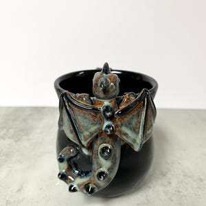 May include: Black ceramic mug with a sculpted dragon handle and body. The dragon features brown, blue, and green accents on its wings and body. The mug has a glossy finish and a unique, fantasy-themed design.