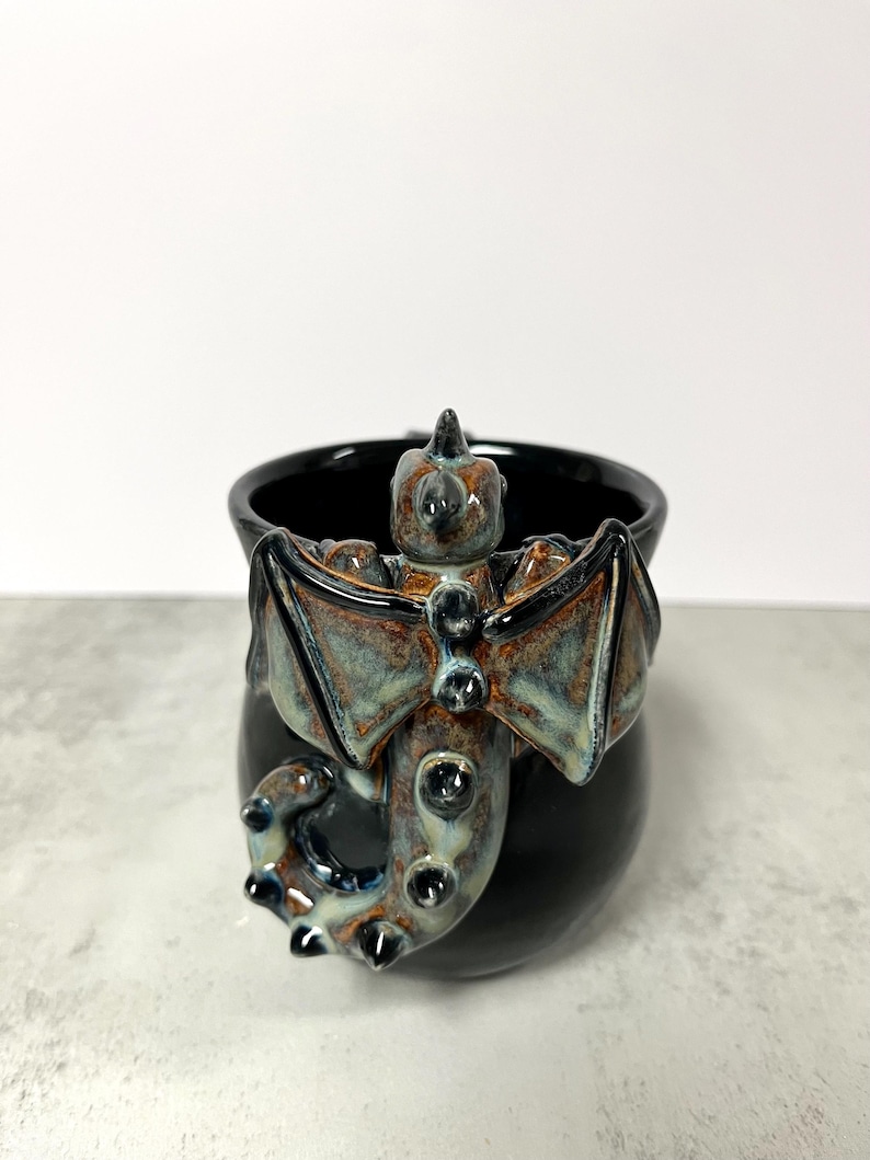 May include: Black ceramic mug with a sculpted dragon handle and body. The dragon features brown, blue, and green accents on its wings and body. The mug has a glossy finish and a unique, fantasy-themed design.